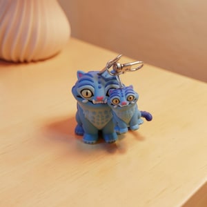 May include: A blue cat keychain with a smaller matching cat charm. The keychains have yellow eyes, pink noses, and white accents. The larger cat is approximately 2 inches tall. The keychain is attached to a silver metal clasp.