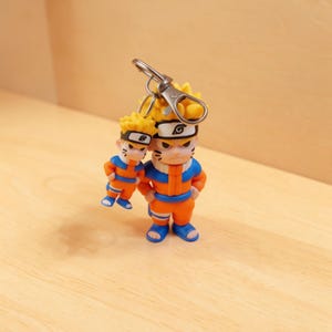 May include: A keychain featuring two Naruto Uzumaki figures in orange and blue outfits with spiky yellow hair and headbands. The larger figure stands upright while the smaller figure hangs from a silver metal lobster clasp and ring attachment.