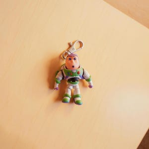 May include: A small figurine of a space ranger character with a purple hood, white suit, green accents, and a silver lobster claw clasp attached to the top. The character has a round face, wide eyes, and a small mouth, with articulated limbs.