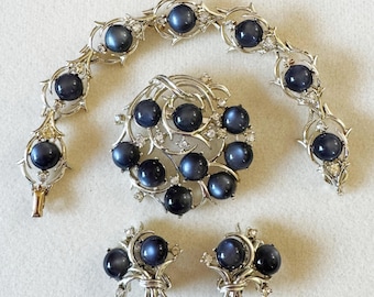 Op de afbeelding: Een zilverkleurige sieradenset met een broche, armband en oorbellen. De set heeft donkerblauwe stenen met een glinsterende afwerking.