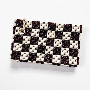 Op de afbeelding: Een rechthoekige clutch met een dambordpatroon. Het ontwerp wisselt af tussen witte en donkerbruine vierkanten, elk versierd met kleine, donkerbruine kralen. Een kleine goudkleurige ketting en ring zijn aan de bovenhoek bevestigd.