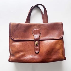 May include: A rectangular brown leather briefcase featuring two top handles, a large front flap, and a central vertical leather strap secured with a brass-toned metal turn-lock closure. The leather displays natural variations in color and texture.