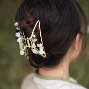 Op de afbeelding: Een gouden haarspeld met een bloemenontwerp met witte bloemen en groene bladeren. De speld heeft de vorm van een driehoek en heeft een veermechanisme.