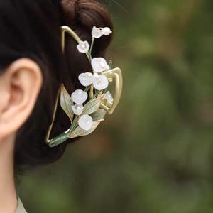 Op de afbeelding: Een gouden haarspeld met een delicaat ontwerp van witte bloemen en groene bladeren. De speld is perfect om elke haarstijl een vleugje elegantie te geven.