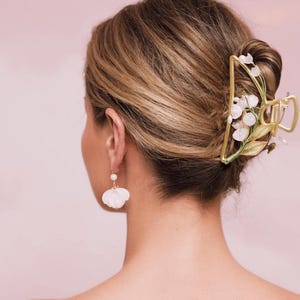 Op de afbeelding: Een gouden haarspeld met witte bloemen en groene bladeren. De speld heeft de vorm van een grote bloem en wordt gebruikt om het haar van een vrouw in een knot te houden.