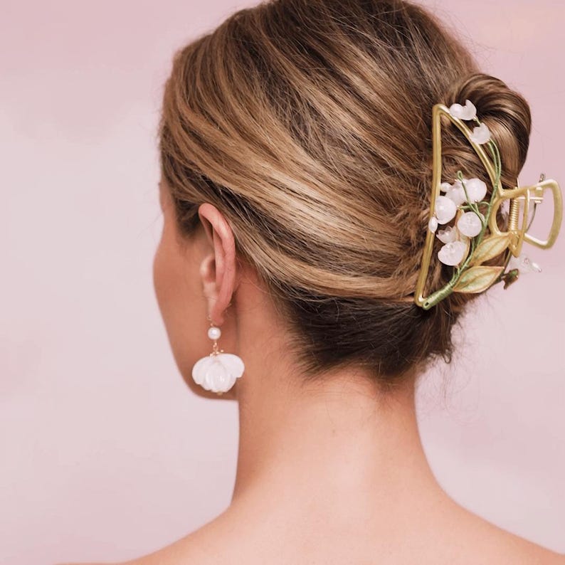 Op de afbeelding: Een gouden haarspeld met witte bloemen en groene bladeren. De speld heeft de vorm van een grote bloem en wordt gebruikt om het haar van een vrouw in een knot te houden.