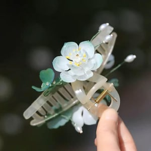 May include: A white flower hair clip with green leaves and pearl accents. The clip is made of plastic and has a spring-loaded mechanism.