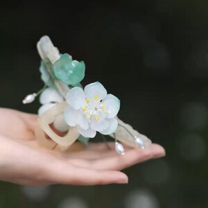 May include: A delicate white flower hair ornament with green leaves and pearl accents. The flower is made of porcelain and has a realistic appearance.