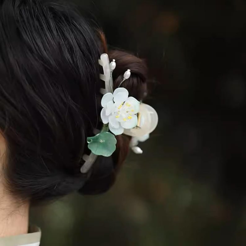May include: A white flower hair clip with green leaves and a white pearl accent. The clip is made of plastic and is designed to hold hair in place.