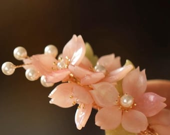 May include: A delicate floral hair accessory featuring translucent pink blossoms and faux pearls. The petals have a subtle sheen, and the piece is accented with small gold details. A beautiful hair clip for special occasions.