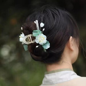 May include: A hair clip with white and green flowers. The clip is made of metal and has a decorative design. The flowers are made of plastic and have a realistic look.