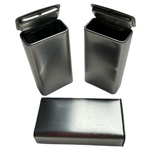 May include: Three brushed metal rectangular containers with hinged lids. Two containers are upright with open lids, and one is lying flat. The containers have a simple, minimalist design and a metallic finish.