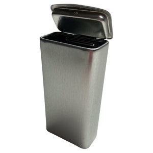 Puede incluir: Una lata de metal plateada con una tapa con bisagras. La tapa está abierta y la lata está vacía.