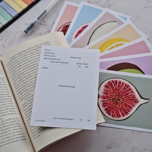 Könnte beinhalten: Eine Sammlung farbenfroher Kunstdrucke mit Früchten, darunter eine Feige, eine Grapefruit und eine Zitrone, sind auf einem Tisch aufgefächert. Ein Buch und ein Notizblock mit Ausfüllfeldern sind ebenfalls vorhanden, zusammen mit einem Stift und Haftnotizen.