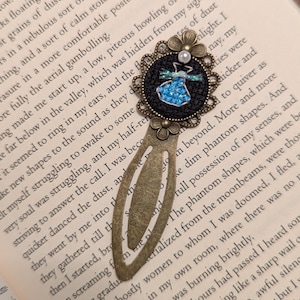 May include: A handcrafted bookmark with a detailed, antique brass finish. The bookmark features a black circular embellishment with a blue beaded design and a pearl accent. The bookmark is resting on the pages of a book.