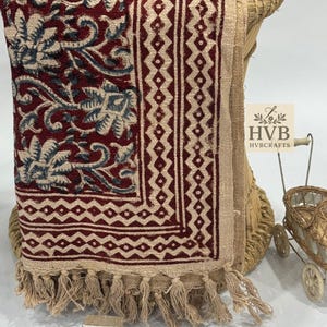 May include: A maroon and beige textile with floral and geometric patterns is draped over a woven chair. The textile has a fringed edge. Two wooden block stamps are in the foreground.