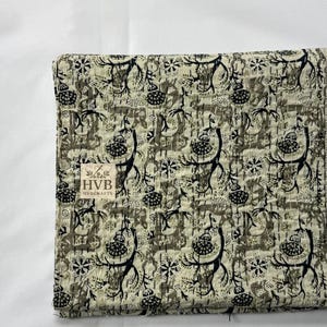 May include: Folded quilt with a repeating pattern of black and olive green floral and abstract shapes on an off-white background. The quilt has a stitched texture and a label that reads "HVB HVBCRAFTS."