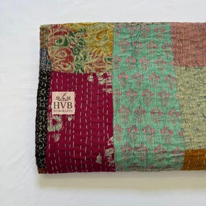 May include: A patchwork quilt with a variety of colors and patterns. The quilt features squares of red, green, yellow, and brown fabric, stitched together with white thread. The quilt has a label that reads "HVB HVBCRAFTS".