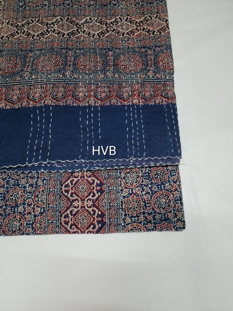 May include: A blue and white kantha quilt with a red and white floral pattern. The quilt is folded in half and the label "HVB" is visible.