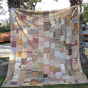 Może przedstawiać: Duża patchworkowa kołdra, wystawiona na zewnątrz, z różnorodnymi kwadratami materiału w odcieniach beżu, różu i brązu. Wzór kołdry jest skomplikowany, z mieszanką wzorów i faktur. Kołdra wykonana jest z mieszanki różnych tkanin.