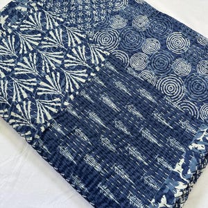 May include: A folded quilt featuring a patchwork design with various shades of blue and white. The fabric displays patterns of white concentric circles, fan-like shapes, and leaf motifs, all held together by visible white running stitch quilting.