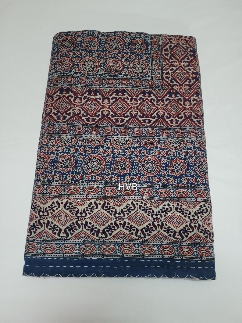 May include: A blue and red patterned cotton quilt with a white background. The quilt has a geometric design and is hand-stitched. The quilt is folded in half and is lying on a white surface. The text "HVB" is visible in the centre of the quilt.