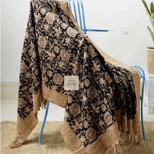 Pode incluir: Uma manta com padrão floral preto e bege drapeada sobre uma cadeira de metal azul. A manta apresenta um design floral repetido e uma borda decorativa com franjas. A manta tem o texto "HVB HANDICRAFTS" em uma etiqueta branca.