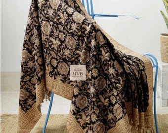 May include: A black and tan floral patterned throw blanket draped over a blue metal chair. The blanket features a repeating floral design and a decorative border with fringe. The blanket has the text "HVB HANDICRAFTS" on a white label.
