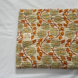 Op de afbeelding: Gevouwen bloemenquilt met een crèmekleurige achtergrond, met een herhalend patroon van grote, gestileerde bloemen in groen- en geeltinten. Bruine stelen en oranje bladeren verbinden de bloemen. De quilt heeft een gestikte textuur en een klein label.