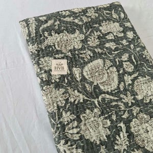 Puede incluir: Un edredón doblado, de color gris oscuro, con un gran estampado floral en blanco roto. El edredón presenta un diseño cosido y una pequeña etiqueta con el texto "HVB MANICRAFTS". El diseño floral incluye grandes flores y hojas.