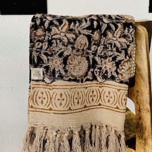 Puede incluir: Una manta plegada en negro y beige con un estampado floral y flecos. La manta está sobre una silla de madera. La manta tiene un borde beige con un diseño repetido y una etiqueta que dice "HVB HANDICRAFTS."