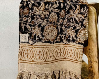 May include: A folded black and tan throw blanket with a floral pattern and fringe. The blanket is draped over a wooden chair. The blanket has a tan border with a repeating design and a tag that reads "HVB HANDICRAFTS."