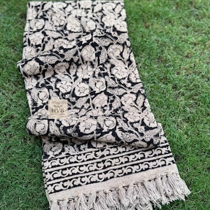 Könnte beinhalten: Ein gefalteter Schal mit schwarzem und beigem Blumenmuster und Fransen. Der Schal hat ein sich wiederholendes Blumenmuster und eine dekorative Bordüre. Das Etikett lautet "HVB HANDICRAFTS".