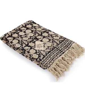Peut inclure: Couverture pliée à motifs floraux noirs et beiges avec des franges. La couverture présente un motif floral répétitif et une bordure décorative. La frange est de couleur beige clair. La couverture est pliée avec soin.