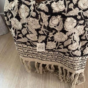 Può includere: Una coperta con motivo floreale nero e beige con frange. La coperta presenta un motivo floreale ripetuto e un bordo decorativo. Le frange sono di colore beige neutro. La coperta è ripiegata in un cesto di vimini.