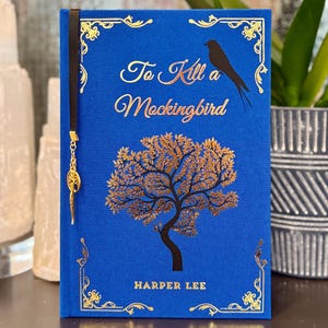 May include: A blue hardcover book with a gold-embossed design. The title is "To Kill a Mockingbird" by Harper Lee. A gold tree and a black bird are featured on the cover.