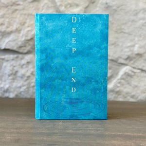 May include: A hardcover book with a teal cover. The title "DEEP END" is printed vertically in white. The cover features a watercolor-like texture and a small illustration of water ripples.