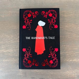 May include: A hardcover book with a black cover featuring a red illustration of a figure in a red cloak and white bonnet. The title "THE HANDMAID'S TALE" is printed in white. Red floral designs frame the image.