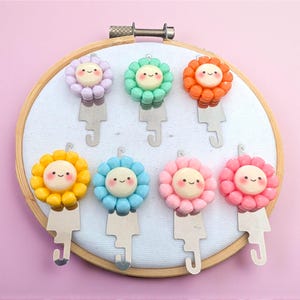 Puede incluir: Una colección de siete llaveros coloridos con forma de flor sonriente. Cada llavero presenta una flor de diferente color, incluyendo morado, verde, naranja, amarillo, azul, rosa y rojo. Los llaveros están unidos a llaveros de metal plateado.