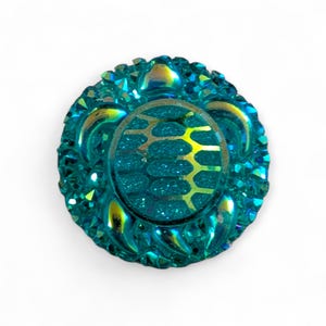 May include: A round, teal-colored button with a glittery center and an ornate, textured border. The center features a gold-colored, honeycomb-like pattern. The button has a glossy, iridescent finish, reflecting shades of yellow and blue.