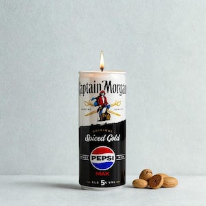 Puede incluir: Una vela hecha de una lata de Captain Morgan Spiced Gold Pepsi Max. La lata es blanca y negra con el logo de Captain Morgan y el logo de Pepsi. La vela está encendida con una llama.