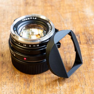 Puede incluir: Un objetivo de c&aacute;mara negro con un anillo plateado y una capucha de lente negra. El objetivo tiene el texto "40mm F 1.4" y "VOIGTLANDER" impreso en &eacute;l.