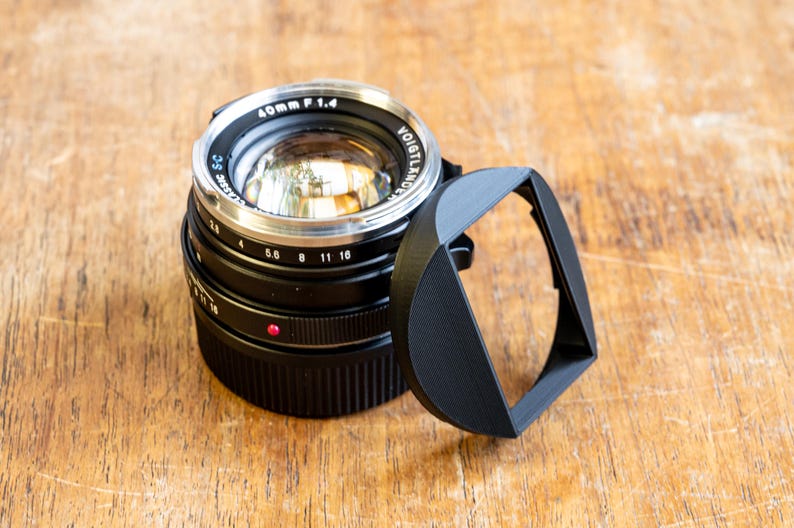 Puede incluir: Un objetivo de c&aacute;mara negro con un anillo plateado y una capucha de lente negra. El objetivo tiene el texto "40mm F 1.4" y "VOIGTLANDER" impreso en &eacute;l.