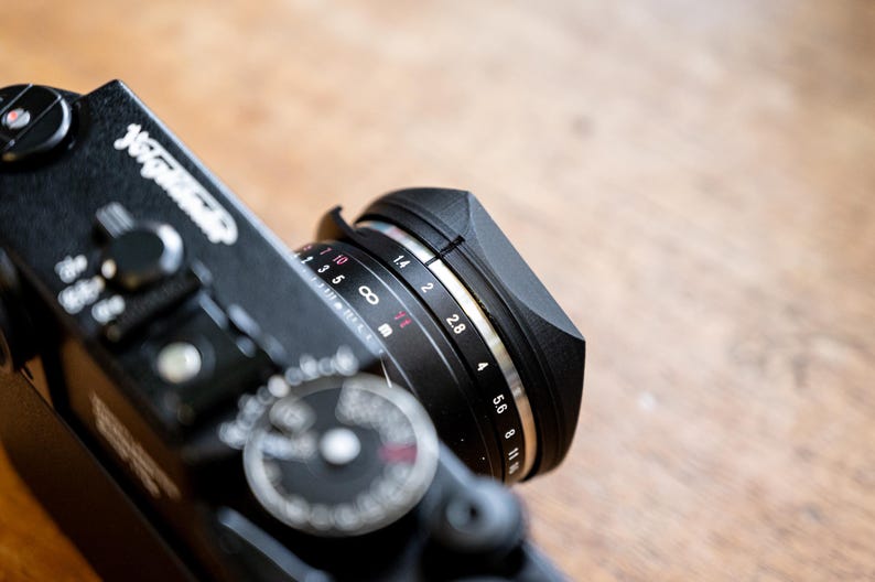 Puede incluir: Una c&aacute;mara negra con un anillo de lente plateado. El anillo de lente tiene marcas para ajustes de apertura de f/1.0 a f/11.