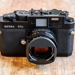 Puede incluir: Una c&aacute;mara de tel&eacute;metro negra BESSA R3A con un objetivo VOIGTLANDER. La c&aacute;mara tiene un dial plateado en la parte superior y un bot&oacute;n plateado en el lateral.