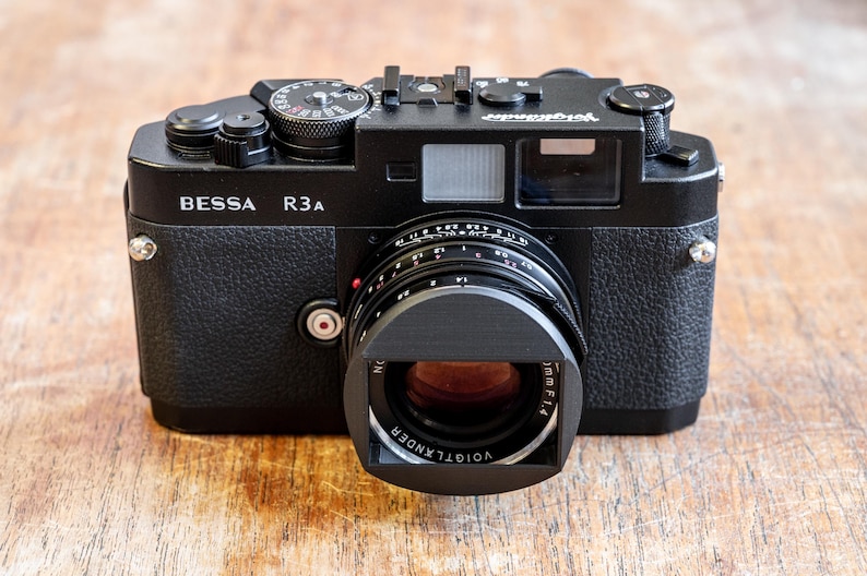 Puede incluir: Una c&aacute;mara de tel&eacute;metro negra BESSA R3A con un objetivo VOIGTLANDER. La c&aacute;mara tiene un dial plateado en la parte superior y un bot&oacute;n plateado en el lateral.