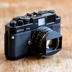 Puede incluir: Una c&aacute;mara de tel&eacute;metro negra BESSA R3A con una tapa de lente. La c&aacute;mara tiene una inscripci&oacute;n plateada que dice "BESSA R3A".