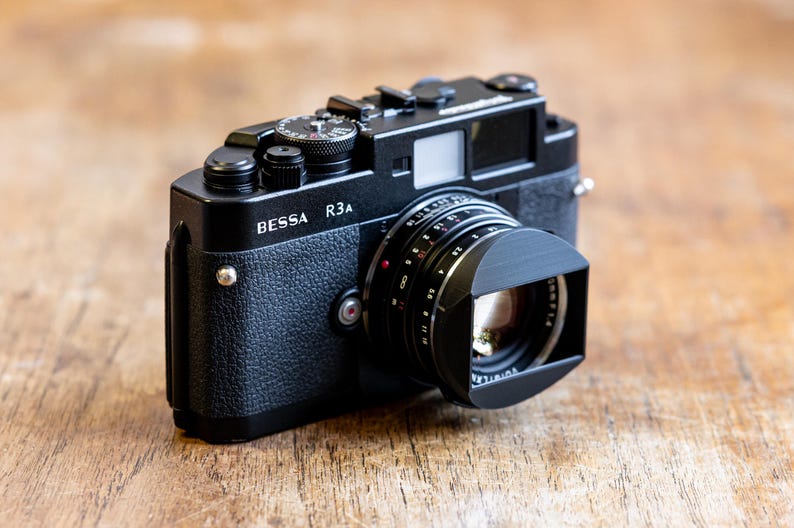 Puede incluir: Una c&aacute;mara de tel&eacute;metro negra BESSA R3A con una tapa de lente. La c&aacute;mara tiene una inscripci&oacute;n plateada que dice "BESSA R3A".