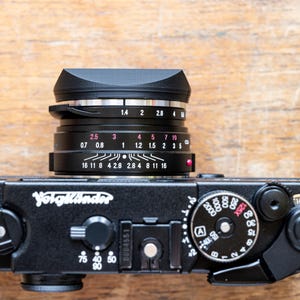 Puede incluir: Una c&aacute;mara Voigtl&auml;nder negra con un anillo de lente plateado y una capucha de lente negra. El objetivo tiene una escala de profundidad de campo con marcas para 0,7, 0,8, 1, 1,2, 1,5, 2, 2,8, 4, 8, 11, 16 y 28. La c&aacute;mara tiene una cubierta de imitaci&oacute;n de cuero negro y un cuerpo de metal negro.