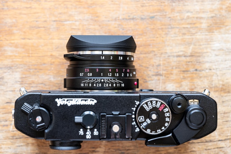 Puede incluir: Una c&aacute;mara Voigtl&auml;nder negra con un anillo de lente plateado y una capucha de lente negra. El objetivo tiene una escala de profundidad de campo con marcas para 0,7, 0,8, 1, 1,2, 1,5, 2, 2,8, 4, 8, 11, 16 y 28. La c&aacute;mara tiene una cubierta de imitaci&oacute;n de cuero negro y un cuerpo de metal negro.